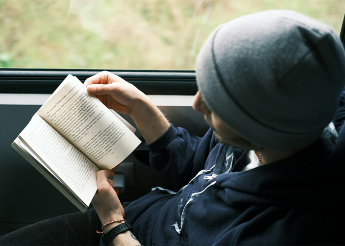 Person wearing a beanie reading a book by a window, illustrating people who still use obsolete things daily.