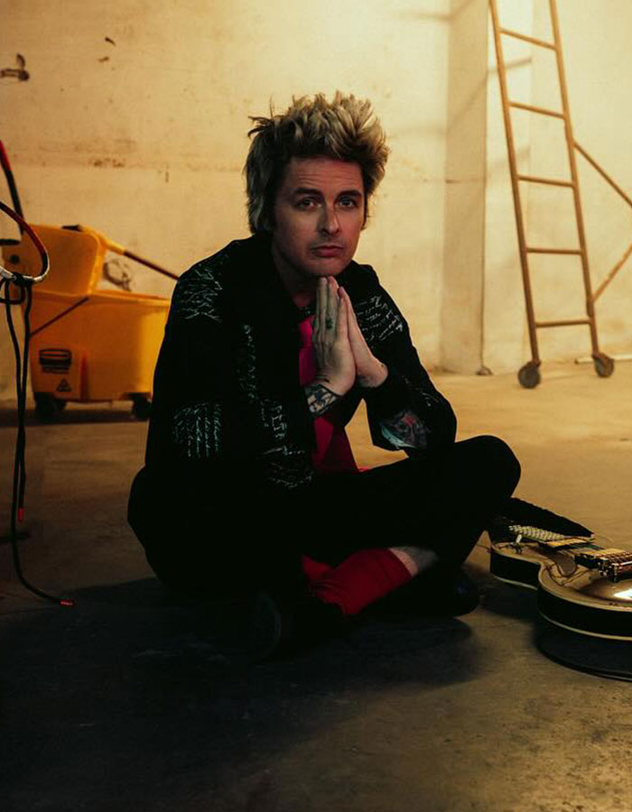 Billie Joe Armstrong of Green Day sitting on floor with guitar nearby in a dimly lit backstage setting.