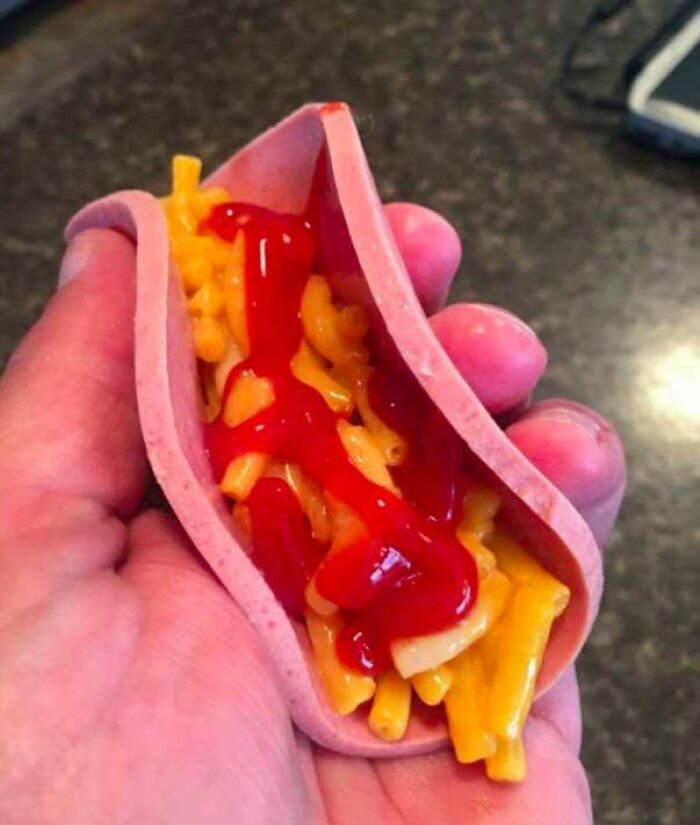 Hand holding a slice of bologna folded with mac and cheese topped with ketchup in a cursed food picture.