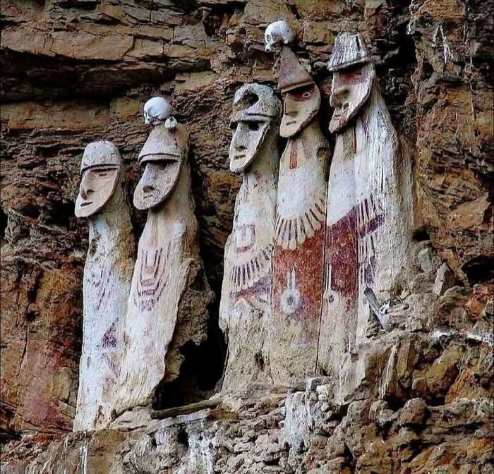 Ancient carved stone figures with painted designs and skulls set against a rocky cliff in archaeology world.