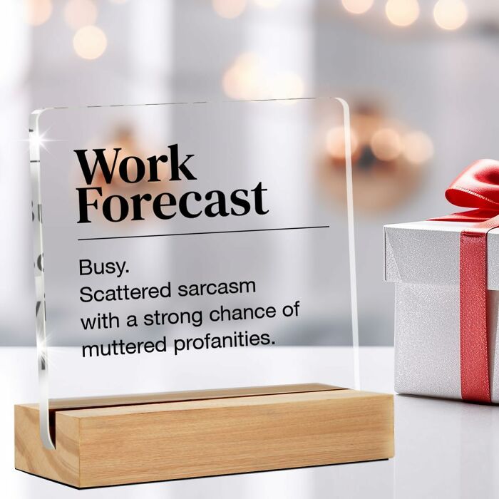 A humorous office supply glass plaque with work forecast text, placed on a wooden stand next to a gift box with a red ribbon.