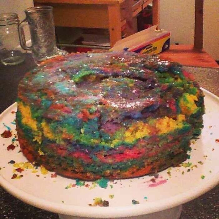 Cursed picture of a colorful, poorly baked rainbow cake with uneven layers and a shiny, sticky surface on a white plate.
