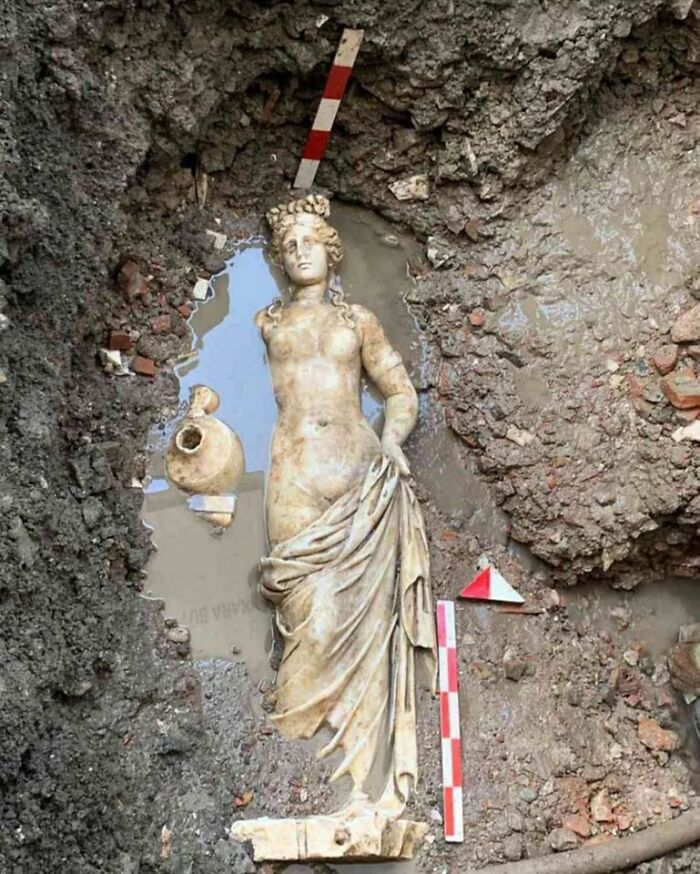 Ancient statue partially buried in archaeological dig site surrounded by soil and measuring tools, archaeology world discovery.