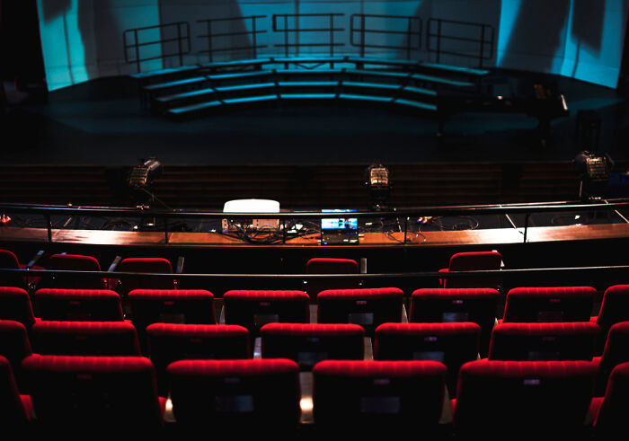 Empty theater seats in front of a dark stage with lighting equipment, illustrating out-of-touch rich people flaunting wealth.
