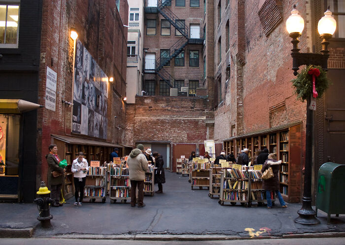 Outdoor urban bookstore with people browsing books against a backdrop of brick walls and vintage street lamps.