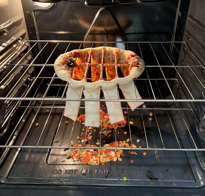 Cursed food picture of a misshapen pizza baking in an oven with toppings spilled on the oven floor.