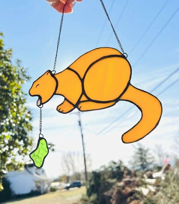 Handmade stained glass craft of an orange cat holding a green fish, an example of horrible crafts shamed by netizens.