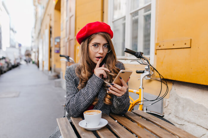 Young woman wearing a red beret, holding a phone with a finger to her lips, hinting at committed lies outdoors.