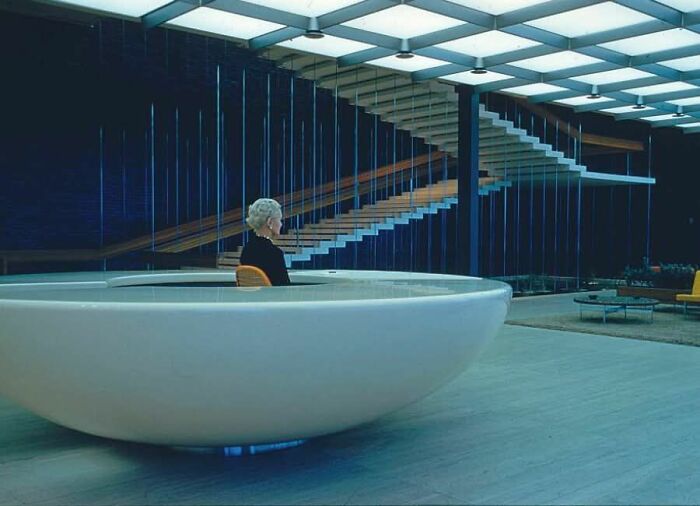 Elderly woman sitting in a modern circular chair inside a futuristic building with floating stairs, historical photos vibe.