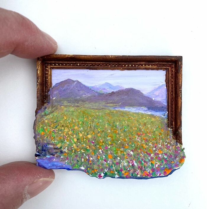 Hand holding a small framed nature scene painting that blends painting and sculpture with flower field and mountains.