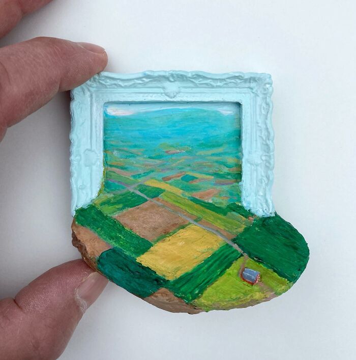 Hand holding a small nature scene artwork blending painting and sculpture with fields and a blue sky.