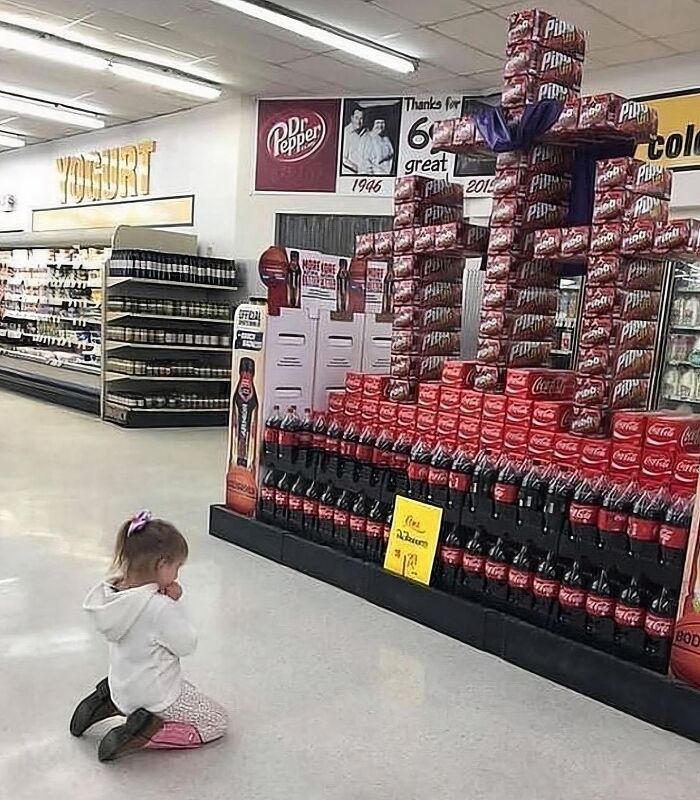 Niña arrodillada en supermercado frente a una figura formada con cajas de refrescos, imagen maldita a todos los niveles.