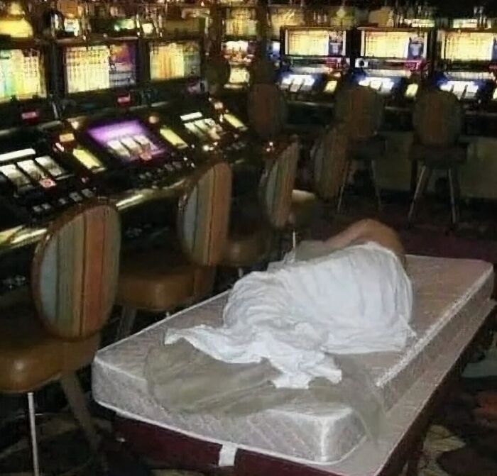 Persona durmiendo en un colchón dentro de casino oscuro entre máquinas tragamonedas, imagen maldita impactante.