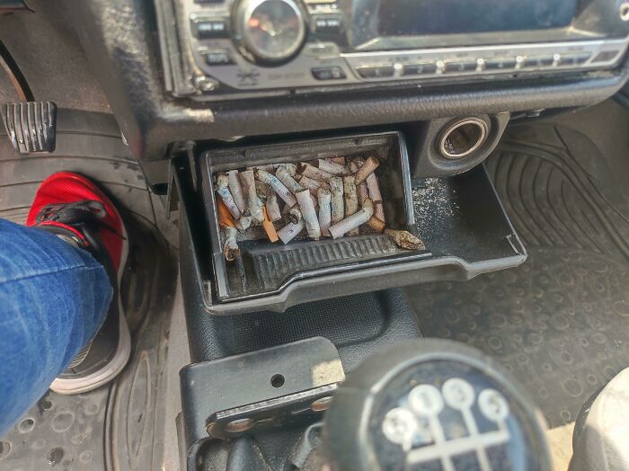 Car ashtray filled with cigarette butts and ashes showing damage in borrowed vehicle interior.