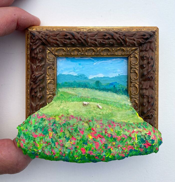 Hand holding a small nature scene combining painting and sculpture with vibrant flowers and rolling hills.