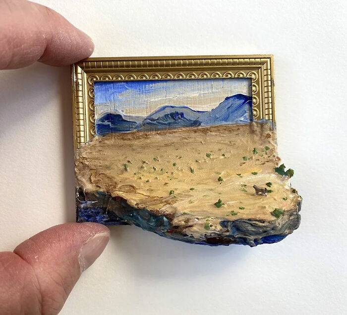 Hand holding a small nature scene combining painting and sculpture with desert and blue mountain elements in a gold frame.