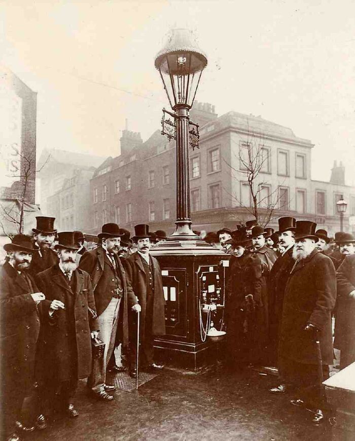 Group of Victorian men and women gathered around an early street telephone box, a strange moment from Victorian times.
