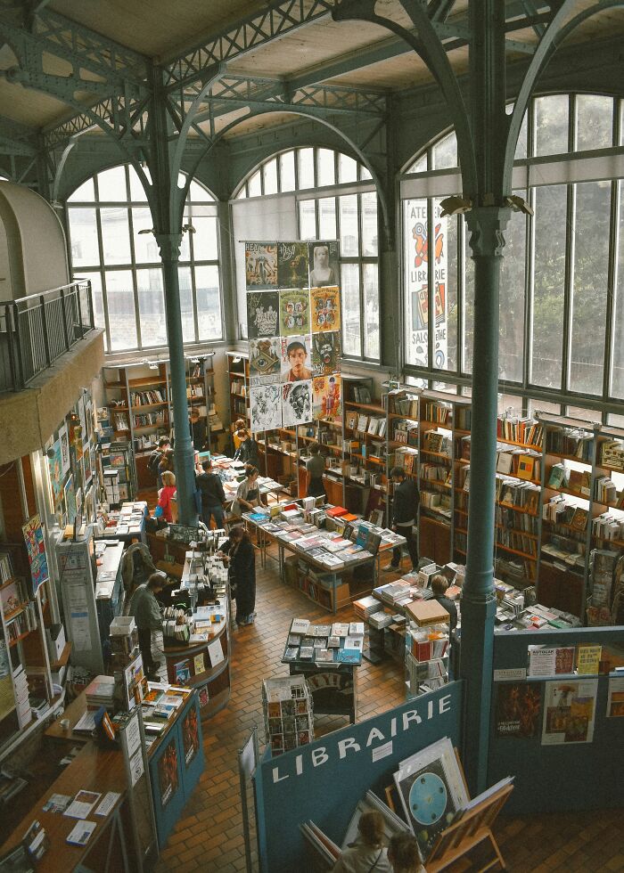 Spacious bookstore with high arched windows and metal beams, filled with books and visitors browsing shelves and tables.