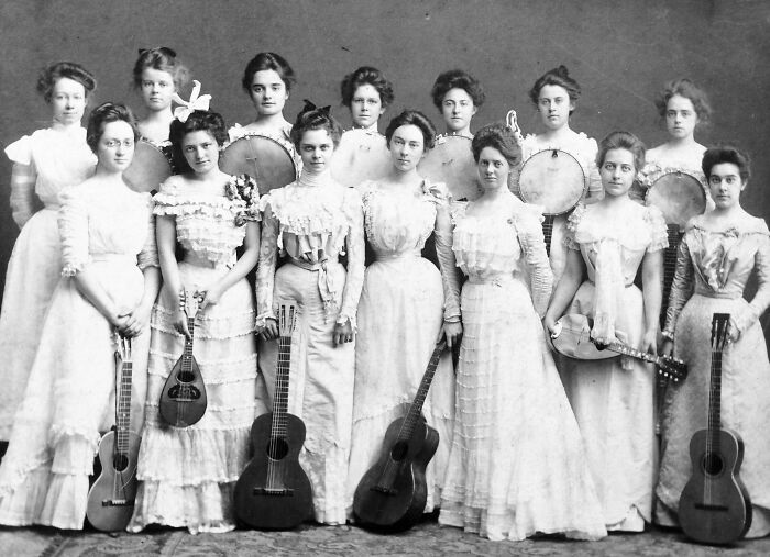 Group of Victorian women dressed in period clothing holding musical instruments in a strange moment from Victorian times.