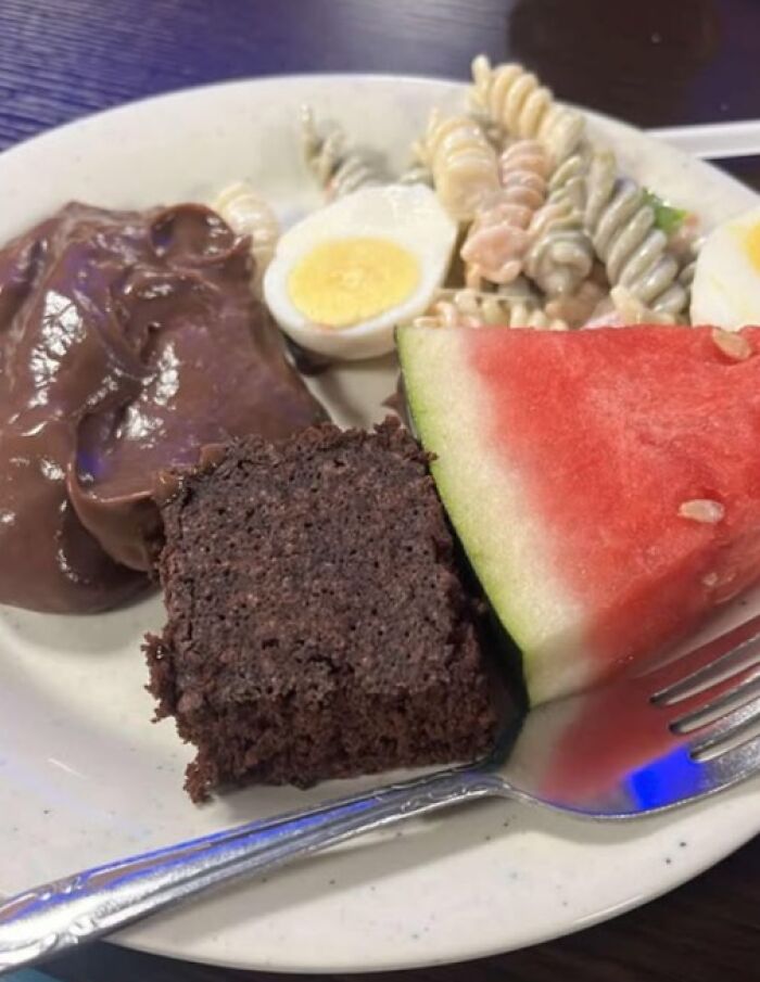 Plate with odd food combination including chocolate pudding, boiled egg, pasta salad, brownie, and watermelon slice, a disgusting food pic.