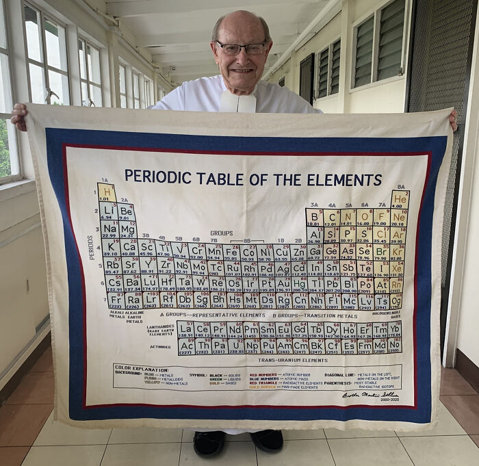 Elderly teacher smiling, holding a large fabric periodic table of elements with detailed chemical information.