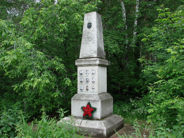 Stone memorial monument with portraits and a red wreath surrounded by dense green forest, evoking unsettling Wikipedia articles.