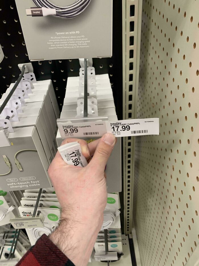 Hand holding a price tag showing rising tariff costs for USB cables on retail store display shelves.
