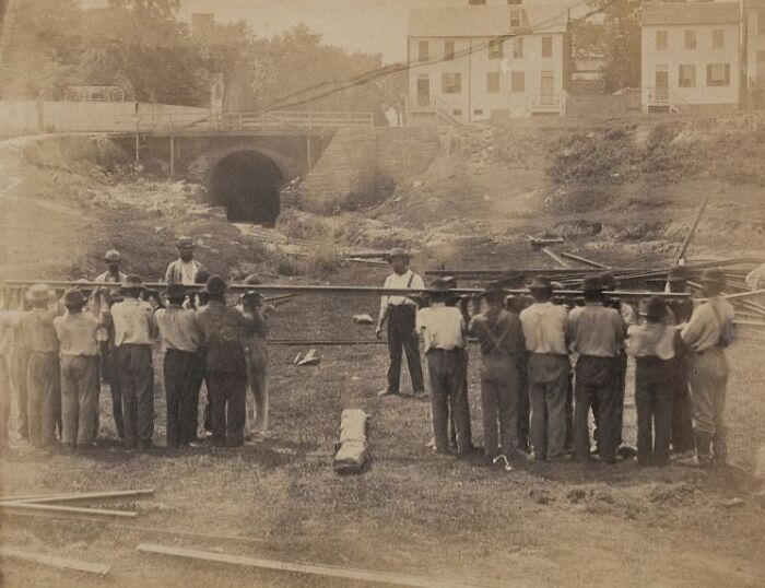 Group of American working class men carrying wooden beams at a construction site in a vintage black and white photo.