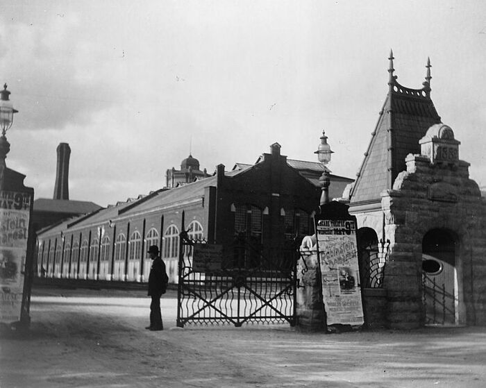 Vintage photo of American working class man standing outside an industrial building near gated entrance in early 20th century.