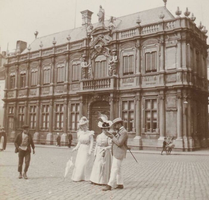 Victorian-era photo showing people in period clothing outside an ornate historic building on a cobblestone street.
