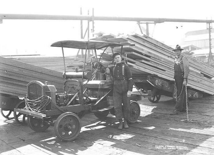 Two vintage American working class men with old truck and large stacks of wooden planks at a job site.