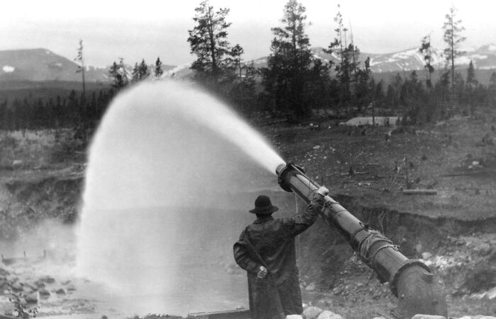Vintage photo of an American working class man operating a large water hose at an outdoor job site with trees and mountains in background