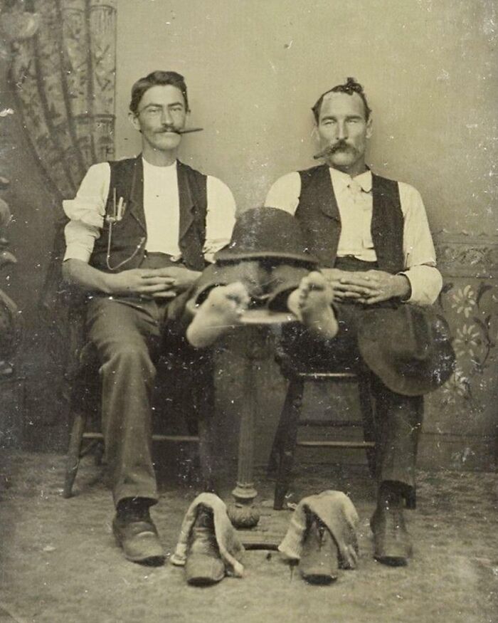 Two Victorian men with mustaches sitting as one appears to balance upside down, a strange moment from Victorian times.