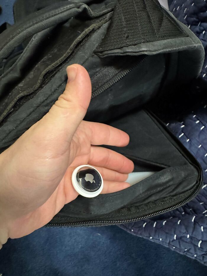 Hand holding an AirTag near the zipper pocket of a black travel bag, illustrating clever travel hacks.