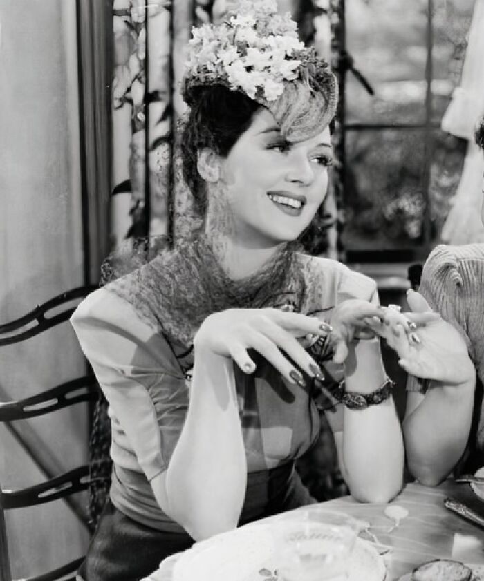 Black and white image of a glamorous 1940s Hollywood icon smiling and wearing a floral hat indoors.