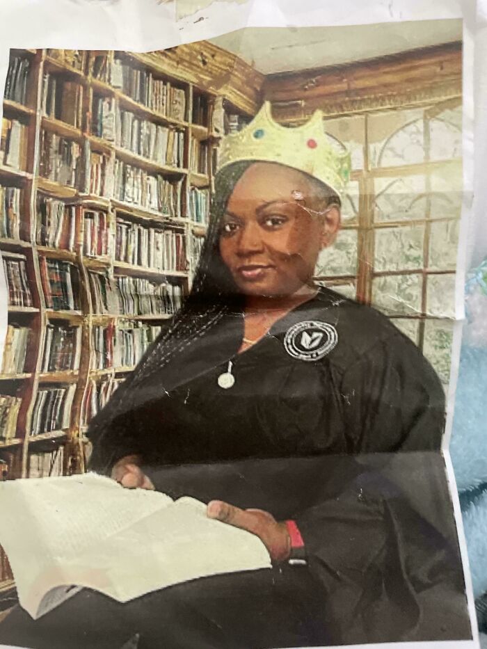 Woman wearing a paper crown and graduation gown sitting with an open book in a library setting in a funny pic.
