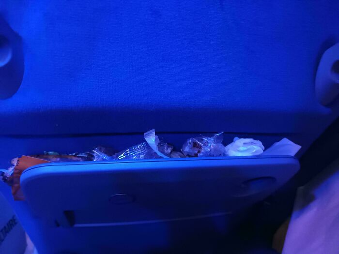 Tray table on a public transport seat cluttered with trash and food wrappers, illustrating frustrating passenger behavior.