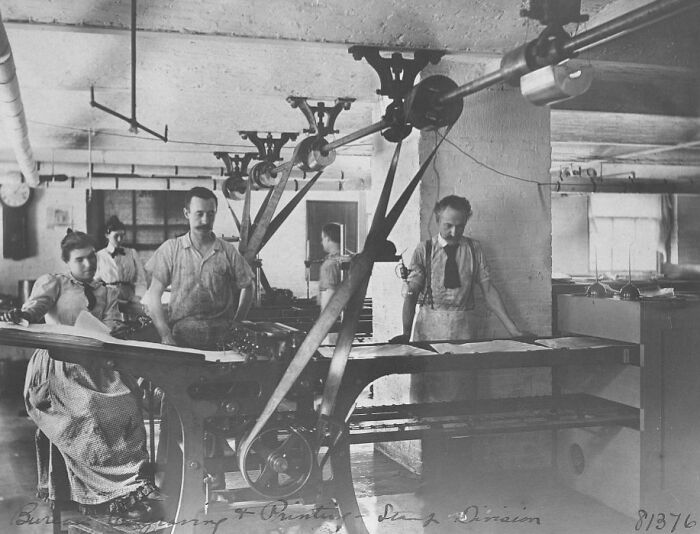 Vintage photo of American working class people operating machinery and handling materials in an early 20th-century factory setting.