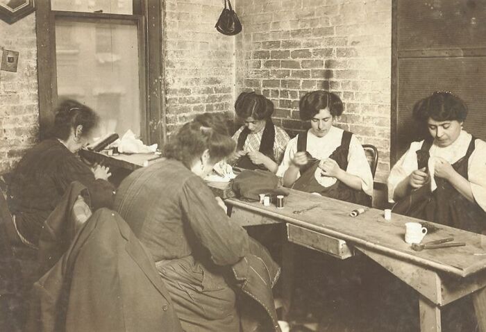 Vintage photo of American working class women sewing garments indoors during their jobs in a brick-walled workshop environment.