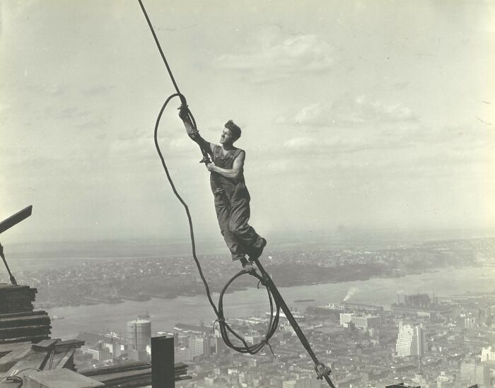 American working class man balancing on steel beam, handling cable high above city during vintage construction work.