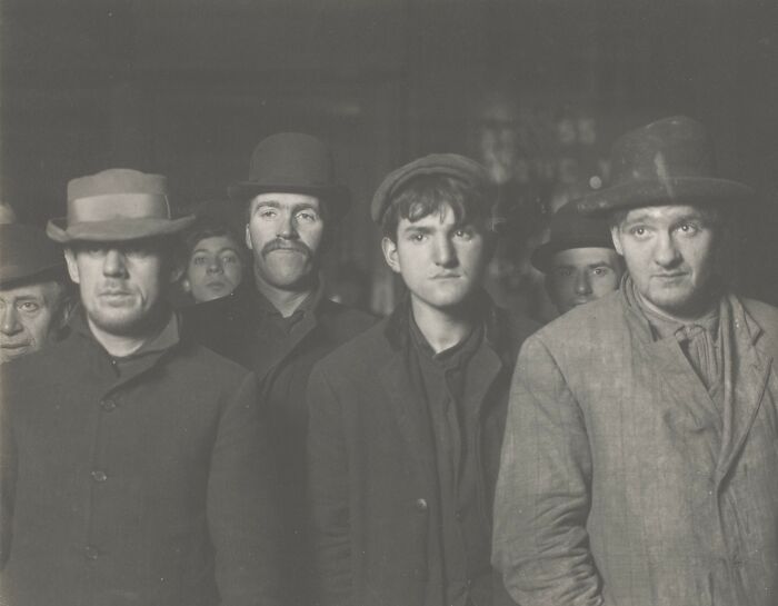 Group of vintage American working class men wearing hats and coats in a black and white photo at worksite.