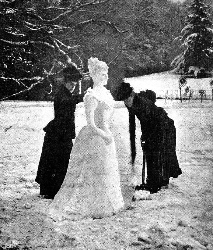 Two Victorian women dressed in black building a strange snow sculpture of a woman in a snow-covered park.