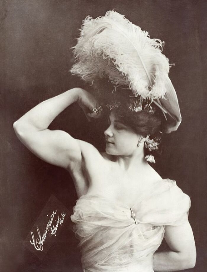Victorian times woman flexing muscles wearing feathered hat and elegant dress in a strange moment from the era