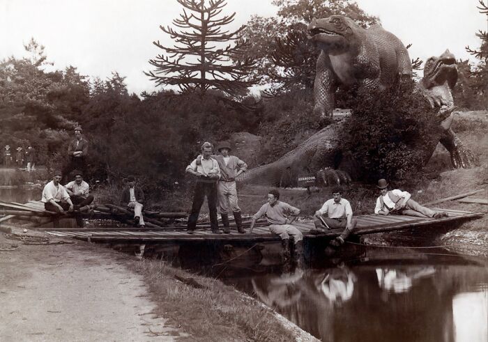 Group of Victorian men by canal near large prehistoric reptile statues, capturing strange moments from Victorian times.