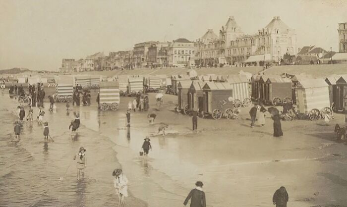 Victorian times beach scene with people in period clothing and bathing huts along the shore in a strange moment.