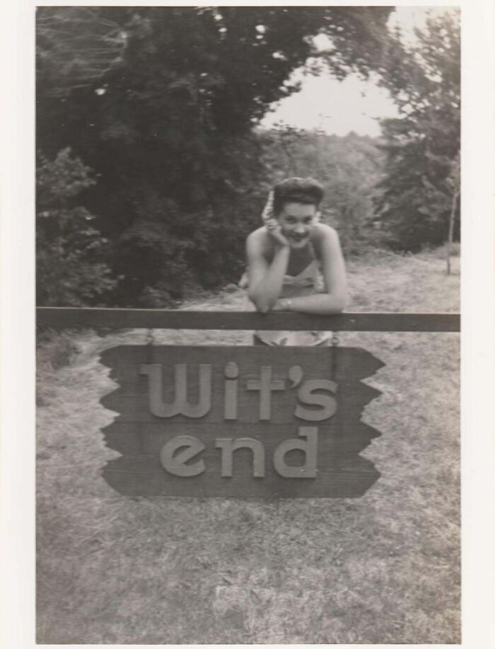 Black and white vintage photo of a woman leaning on a wooden fence with a sign that reads Wit's End outdoors.