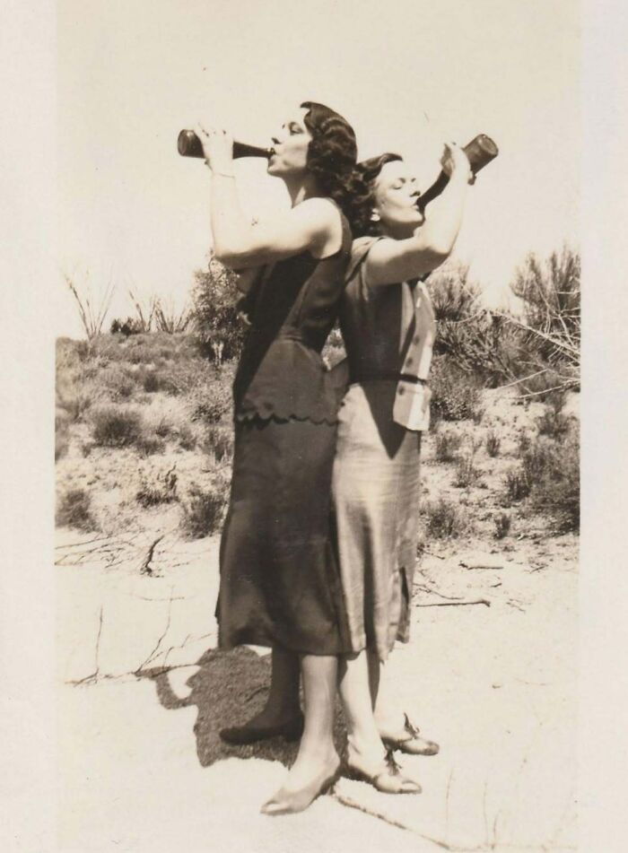 Two women in vintage dresses drinking from bottles back-to-back in an outdoor setting, vintage photo with historical charm.