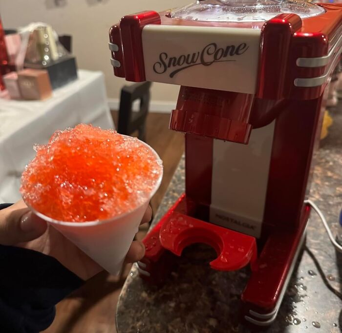 Red and white snow cone machine with a hand holding a cup of bright orange shaved ice, a quirky kitchen gadget.