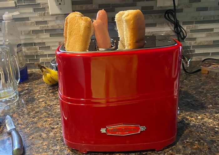 Red kitchen gadget toaster designed for hot dogs with buns on a granite countertop and modern backsplash tiles.