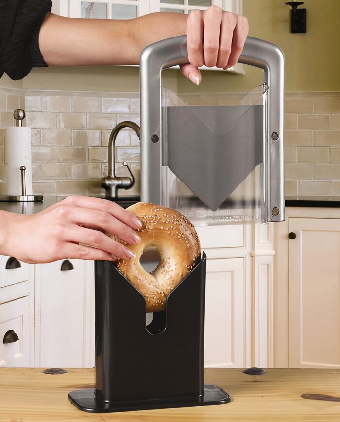 Hand slicing a bagel using a unique kitchen gadget in a modern kitchen counter setting with beige tiles.